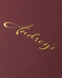 Gold metallic embossed cursive typography reading Audreys on a solid dark burgundy textured paper surface