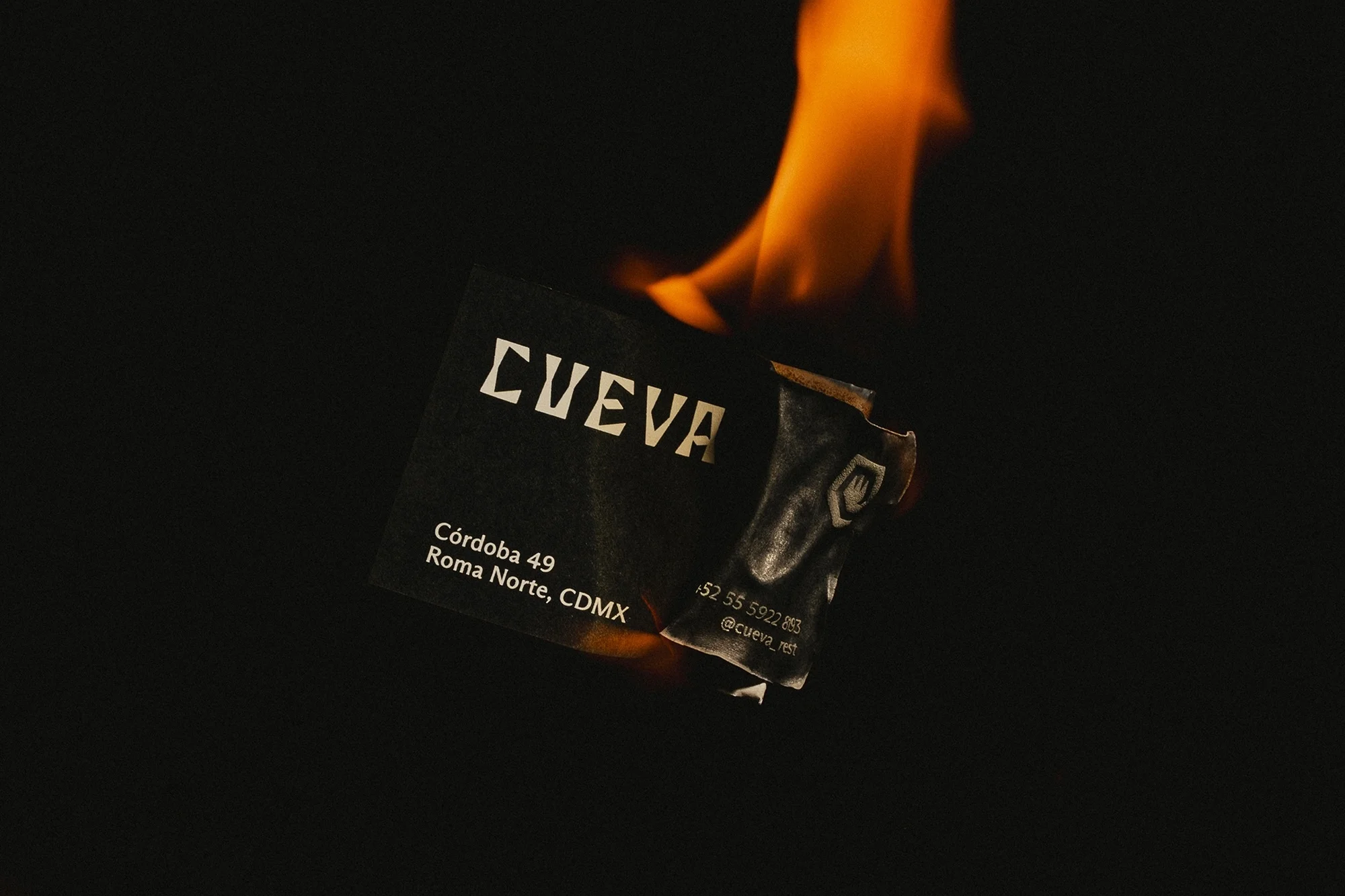 A partially burned business card for 'Cueva' is captured against a black background with a vibrant orange flame licking its surface.