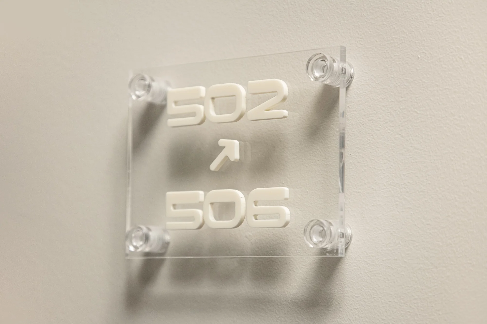 Office directional sign on a white wall indicating room numbers 502 and 506. Clear acrylic with white lettering and arrow.
