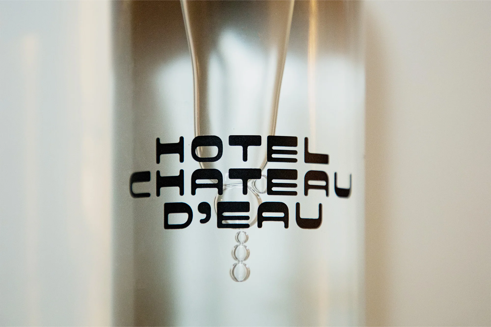 Close-up shot of a transparent glass water dispenser with a black 'Hotel Chateau D'Eau' logo on it against a clean white background.