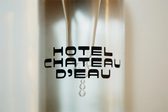 Close-up shot of a transparent glass water dispenser with a black 'Hotel Chateau D'Eau' logo on it against a clean white background.