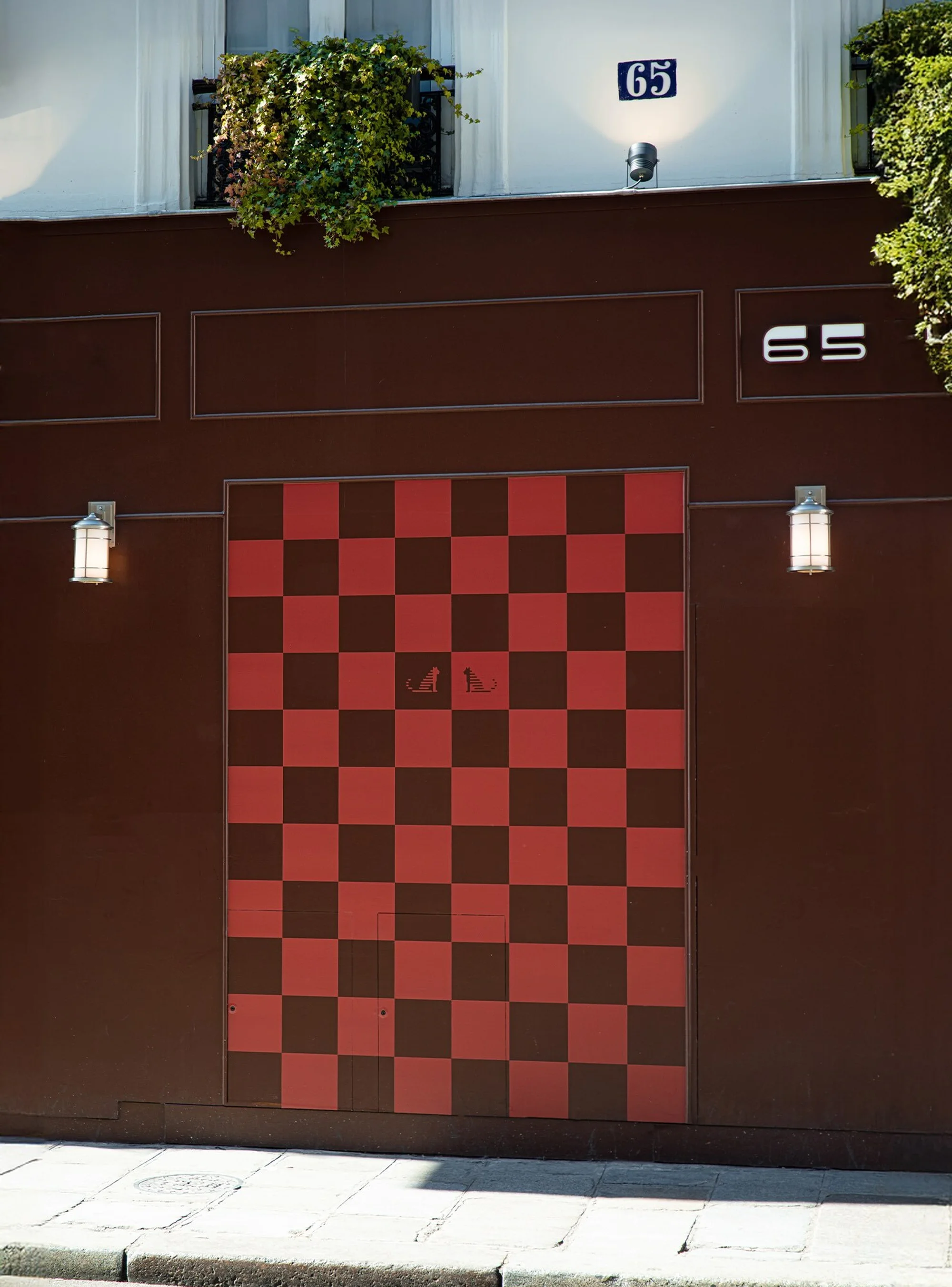 Building facade with a checkered pattern door, a window box with plants above, and a street view in the foreground on a sunny day.
