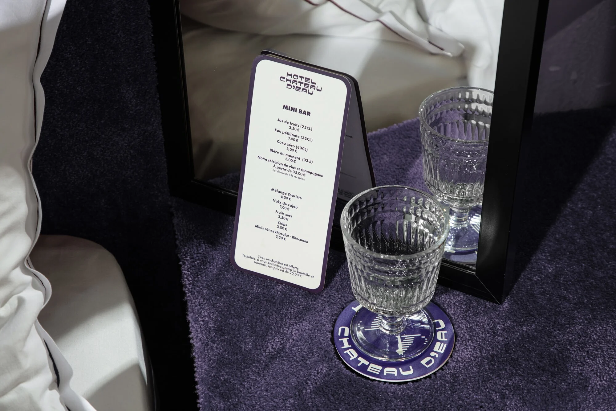 Hotel mini bar menu next to two clear textured glasses on a purple surface with bed linens in the background.