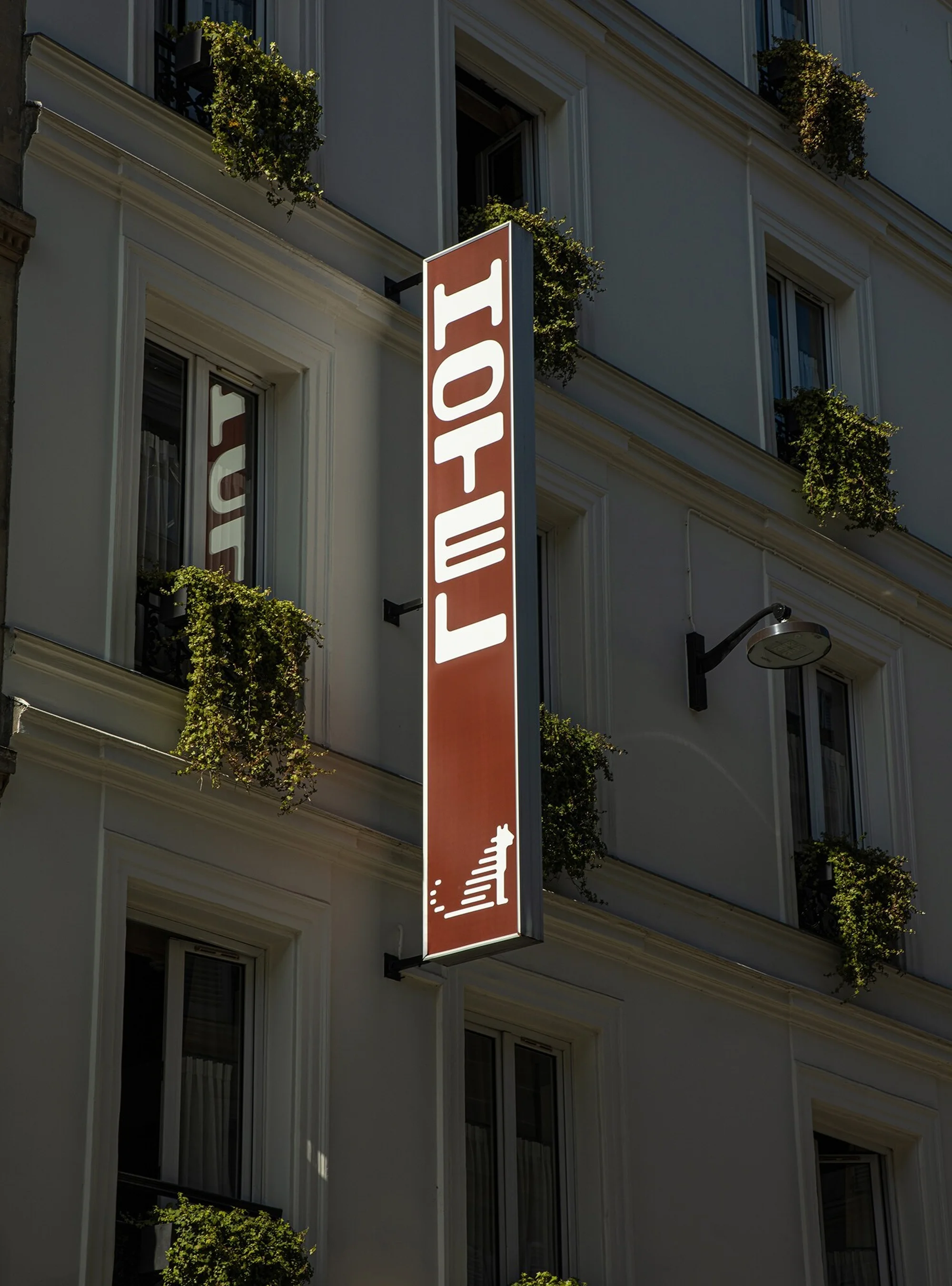 Facade of a white building with multiple windows and a vertical 'HOTEL' sign. Greenery in window boxes adds a touch of nature.
