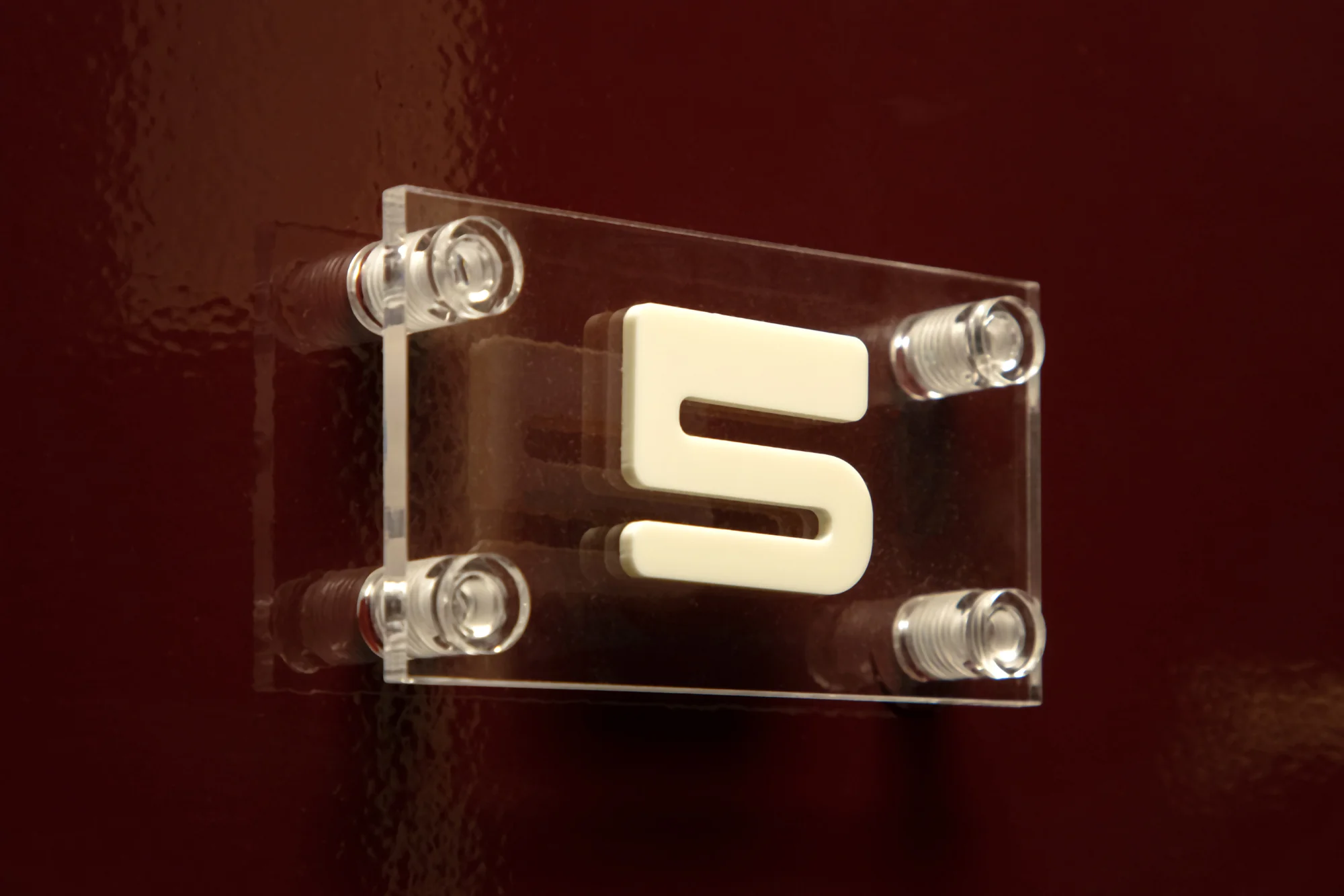 Close-up of clear acrylic sign with off-white '5' attached to a deep red, glossy wall, secured with clear acrylic bolts.