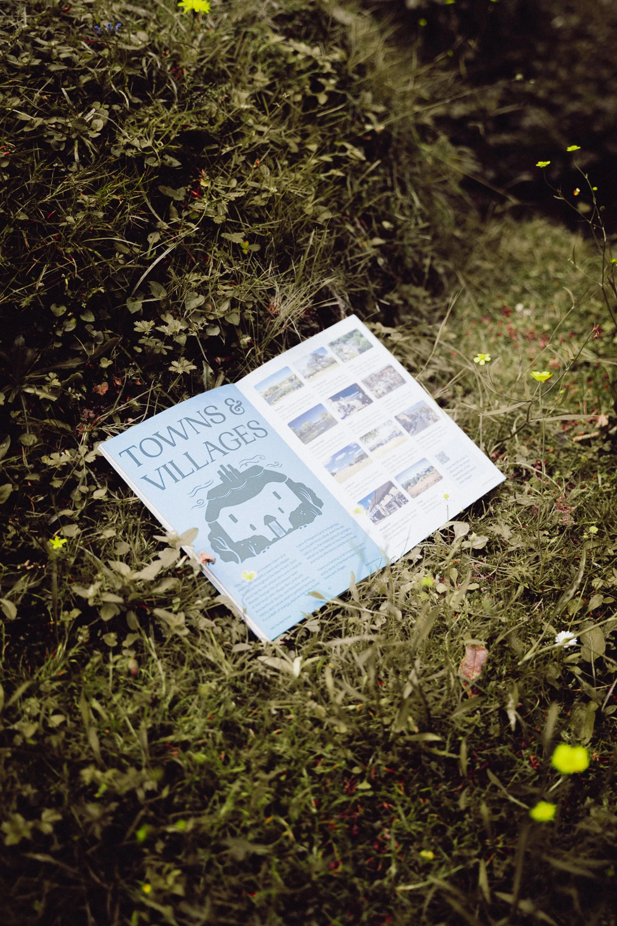 Open travel brochure titled 'Towns & Villages' sits on green grass with small yellow flowers in an outdoor, natural environment.