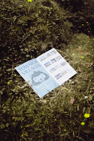 Open travel brochure titled 'Towns & Villages' sits on green grass with small yellow flowers in an outdoor, natural environment.