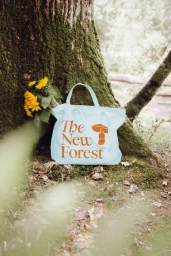 Light blue canvas tote bag with brown text resting against a large tree trunk next to yellow sunflowers