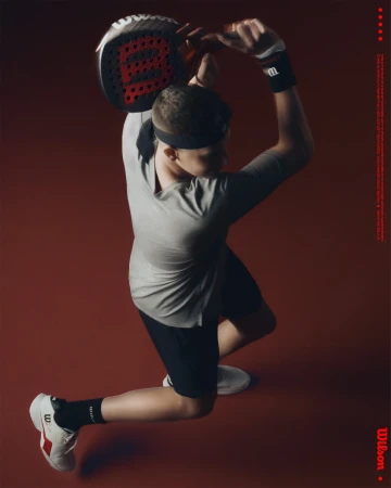 Male padel player in gray tee and black shorts, prepares to strike the ball in a dynamic pose against a solid red backdrop.