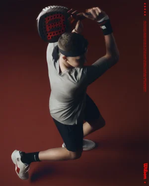 Athlete holding a padel racket overhead in a dynamic swinging pose against a dark reddish-brown background.