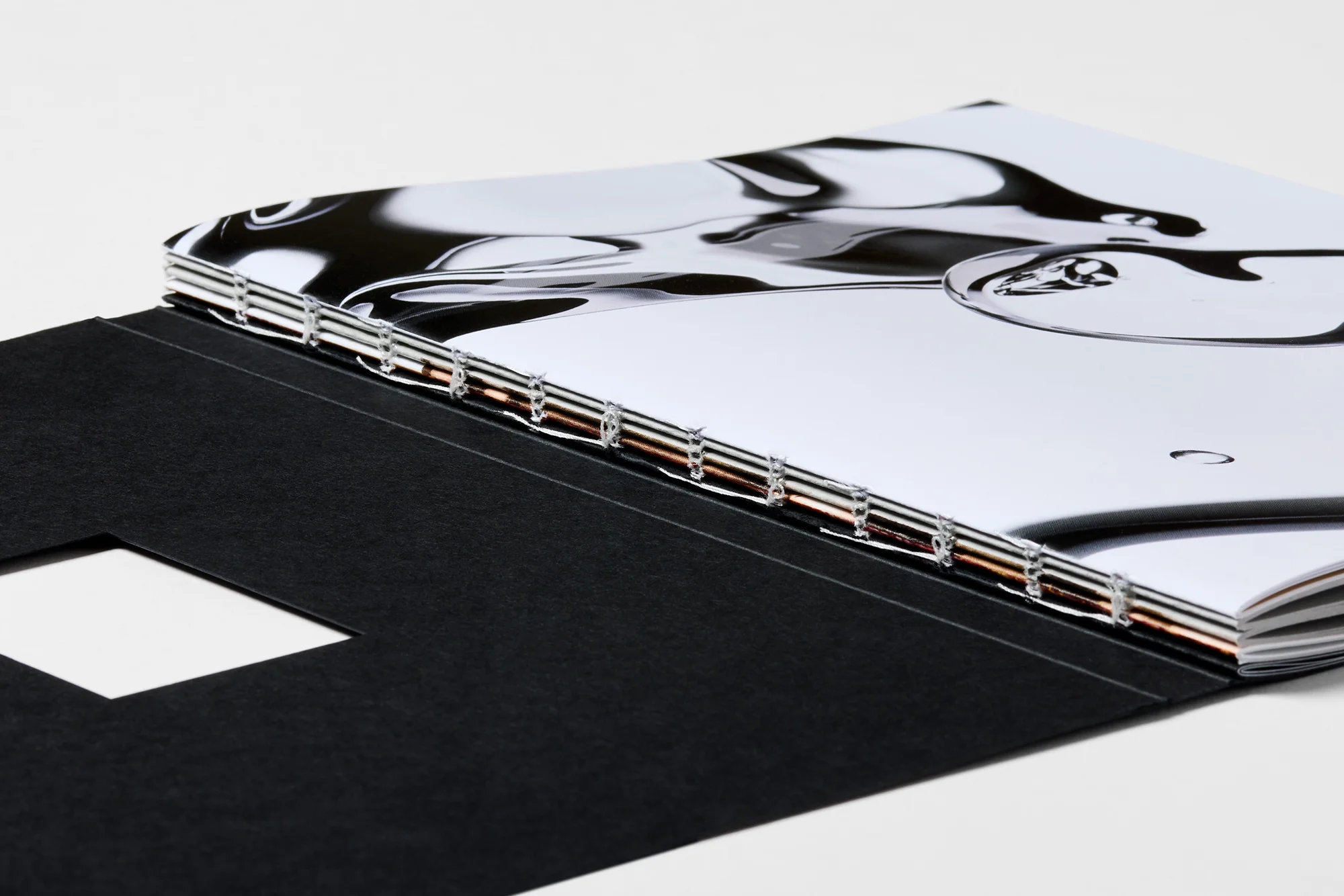 Close-up of an open book or magazine with a dark cover featuring a die-cut square, showcasing its binding and a glossy page.