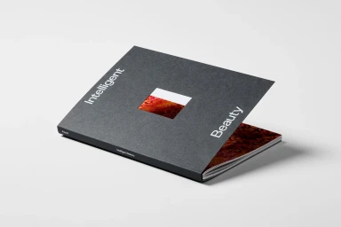 A dark grey booklet titled Intelligent Beauty resting on a clean white surface with a square cutout revealing a reddish image inside
