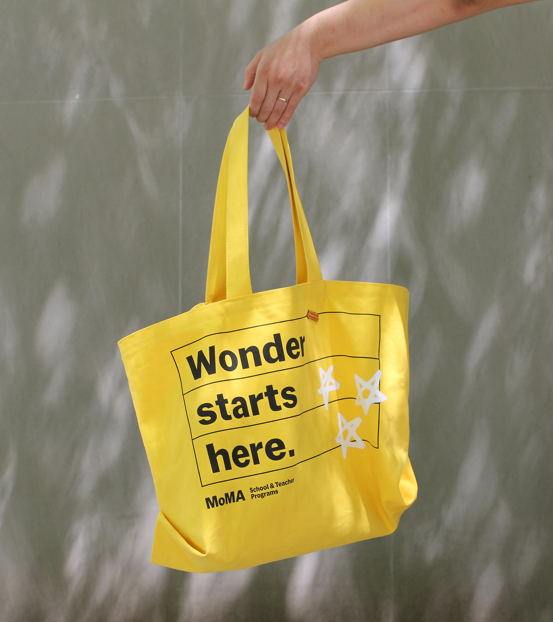 Person holding a bright yellow tote bag with black lettering and white star graphics against a gray wall with shadows.