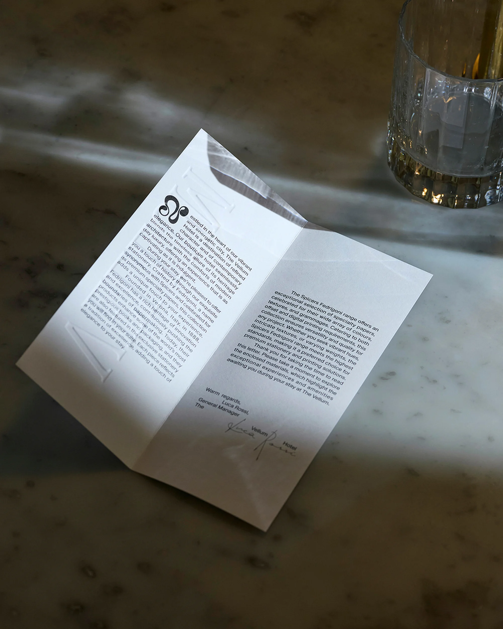 Brochure sits on a marble surface with a patterned glass nearby. The brochure is open, displaying text on both sides of the opened paper.