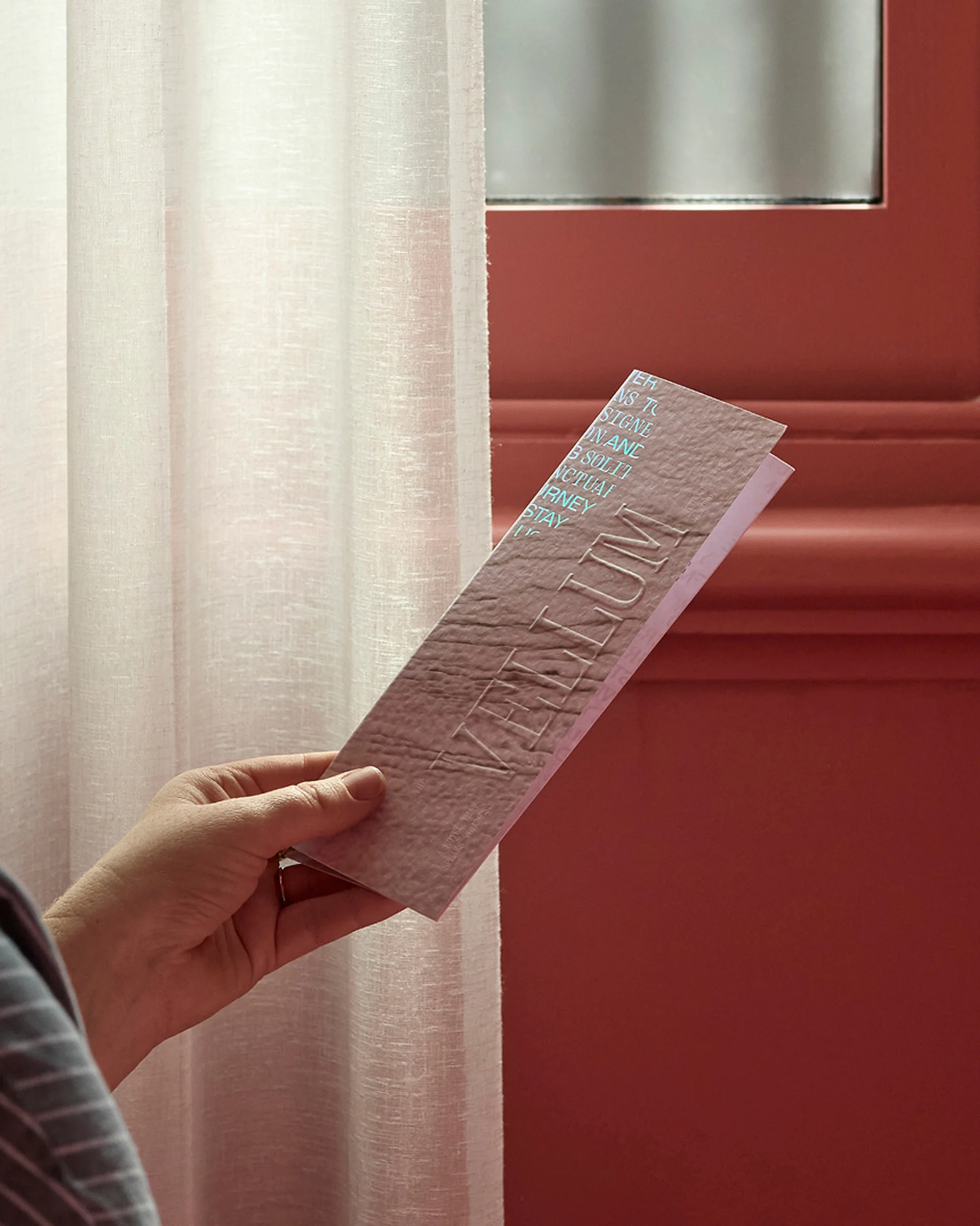 A woman's hand holds a promotional paper printed with 'Vellum' in embossed lettering, set against a salmon-colored wall and sheer white curtains.