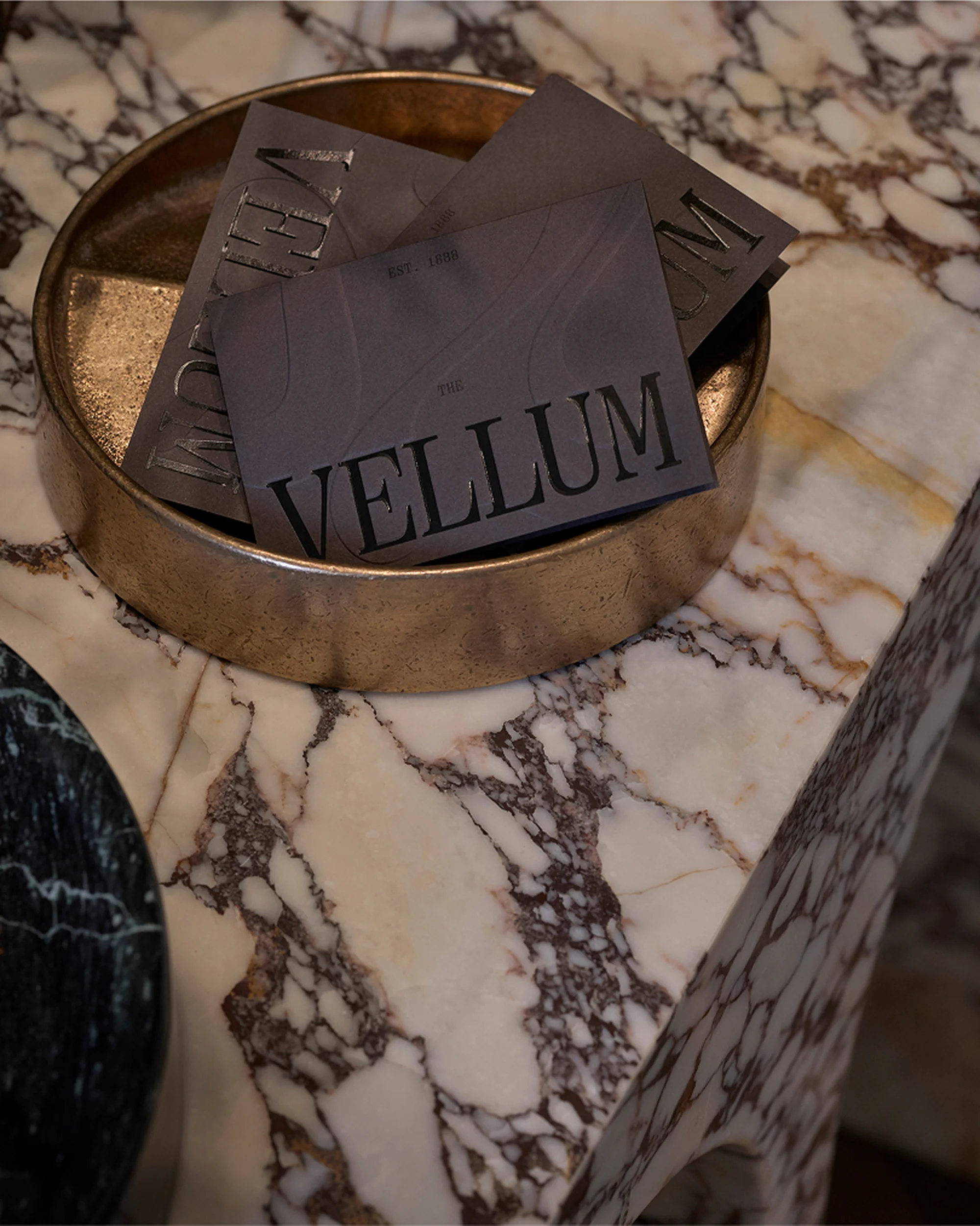 Three dark business cards with the word 'VELLUM' rest inside a shallow bronze bowl on a white marble table with dark veining.