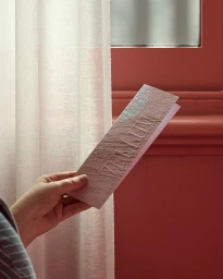 Hand holding a textured pink paper brochure with debossed typography against a sheer curtain and terracotta wall
