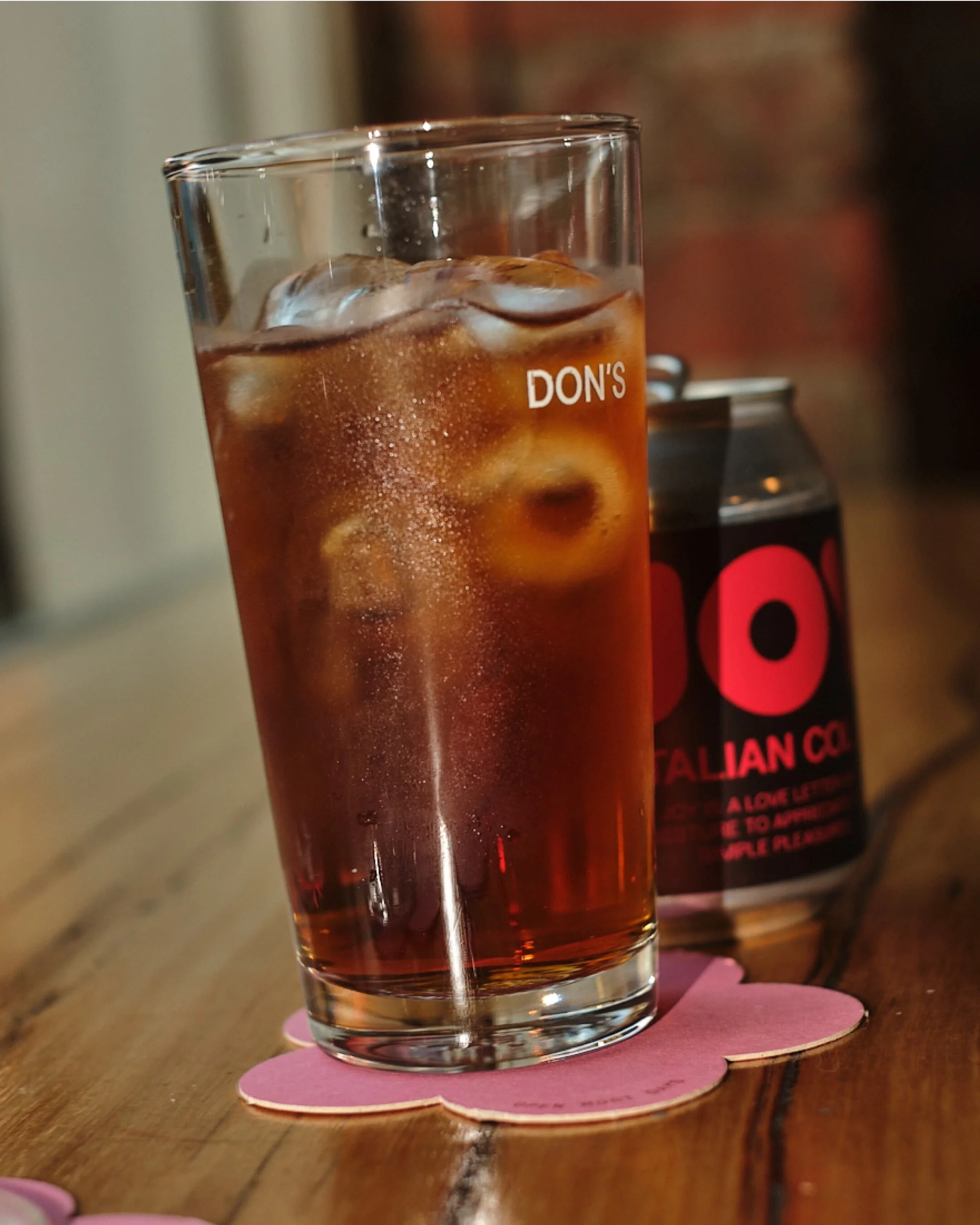 Tall glass of iced drink with lemon peel next to soda can on a pink flower coaster, presented on a wooden surface, indoor setting.