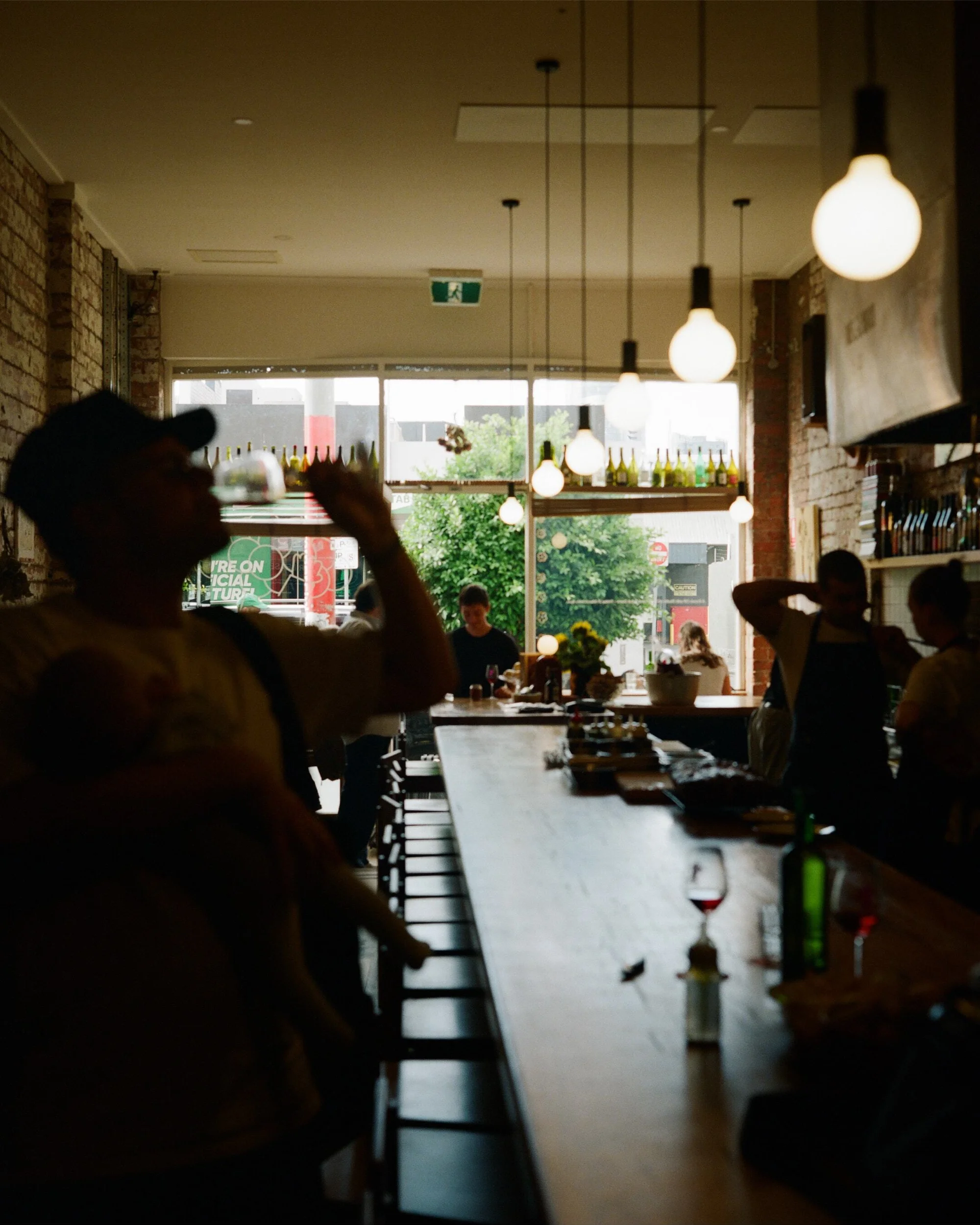 People at a bar with exposed brick walls, pendant lighting, and large window showing outdoor greenery in an urban environment.