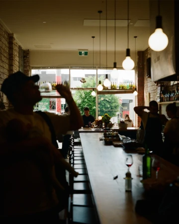 People at a bar with exposed brick walls, pendant lighting, and large window showing outdoor greenery in an urban environment.