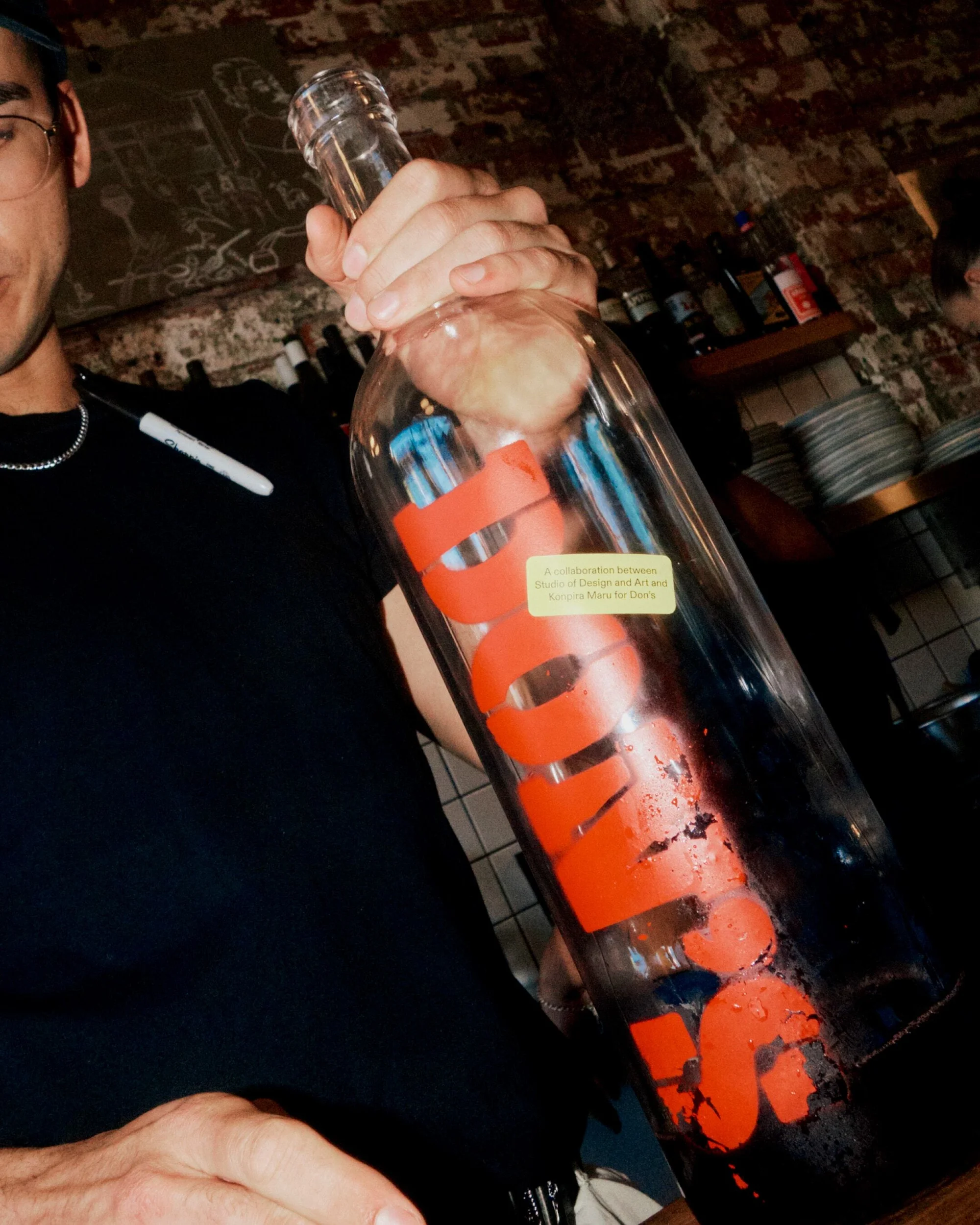 Adult male holding large glass bottle with red painted logo inside, standing in bar, wearing black shirt and glasses.