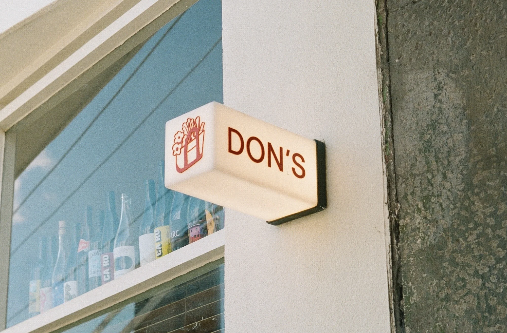 Outdoor daytime view of a white rectangular illuminated sign reading DON'S, affixed to a building exterior with a window in the background.