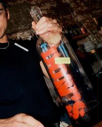 Close-up of a person holding a large glass bottle with bold red Don's branding in a bar setting