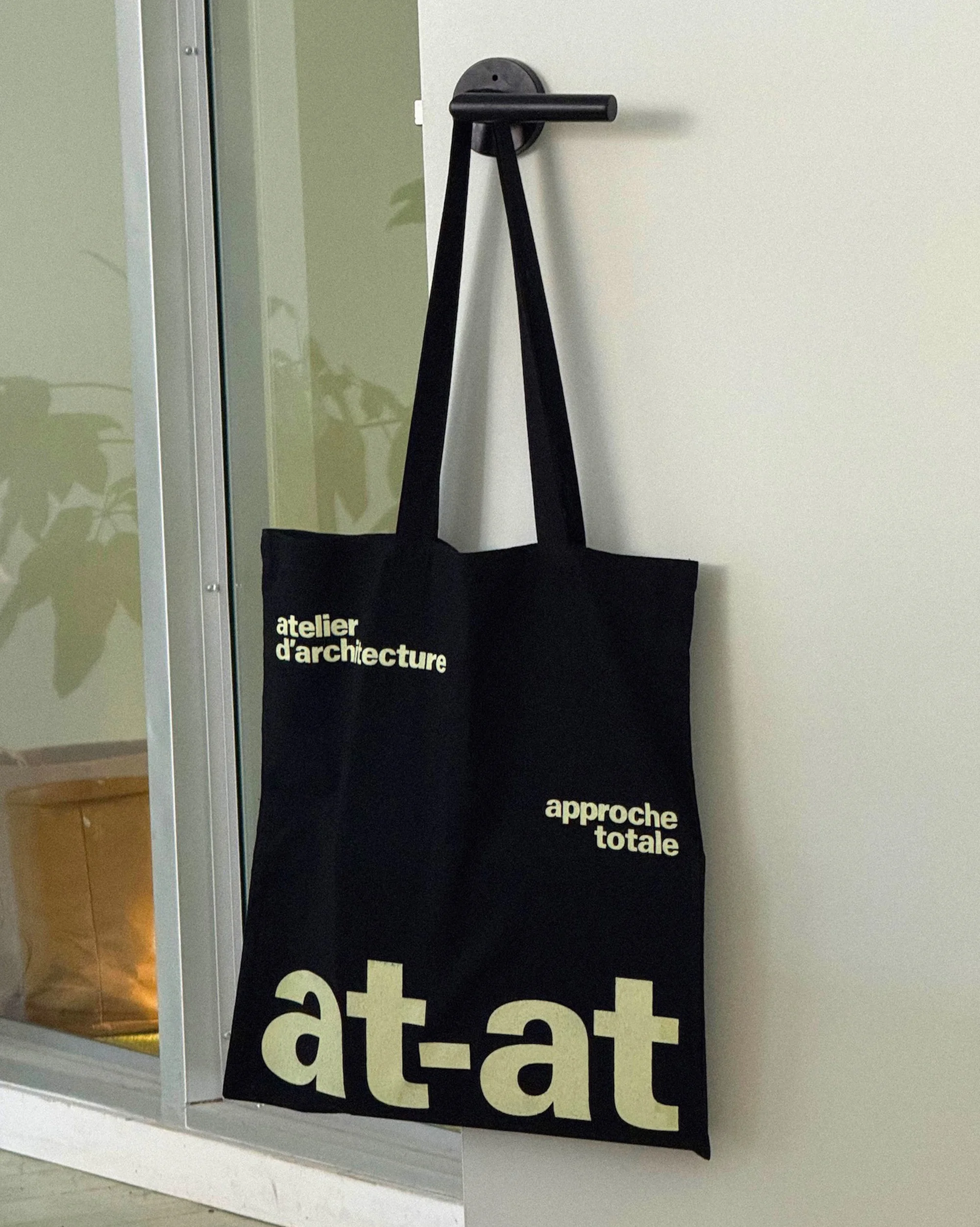 Black tote bag hanging on a door handle, featuring 'atelier d'architecture' and 'approche totale' text, against a white wall indoors.