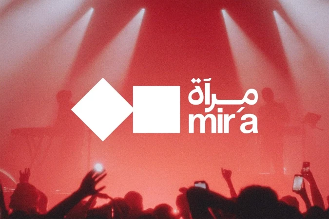 Concert crowd with raised hands and cell phones in front of band on stage, bathed in red stage lights and a logo projected above.
