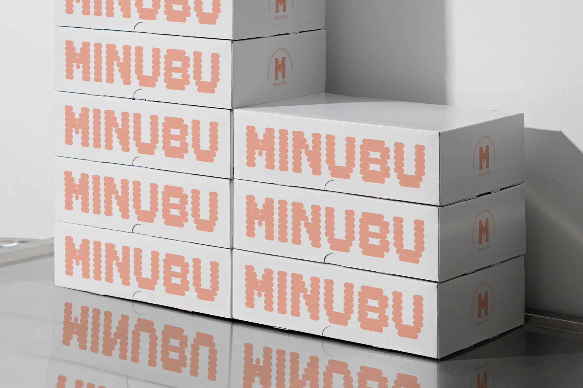 Stack of white product boxes with pink pixelated 'MINUBU' logo against a neutral wall and surface in a studio setting.
