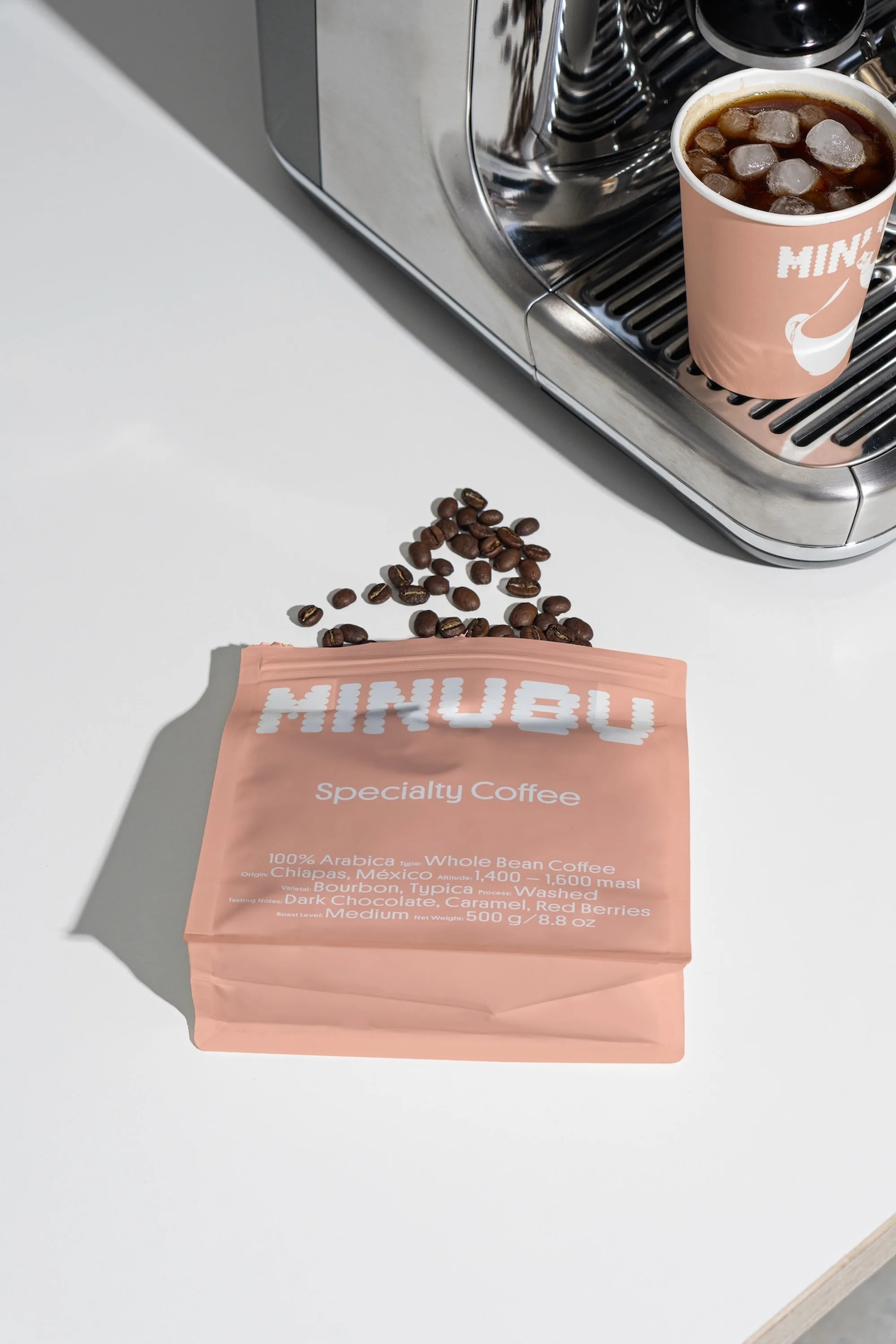 Close-up of Minubu specialty coffee bag with coffee beans spilling out, set next to a coffee maker and iced coffee in a pink cup.