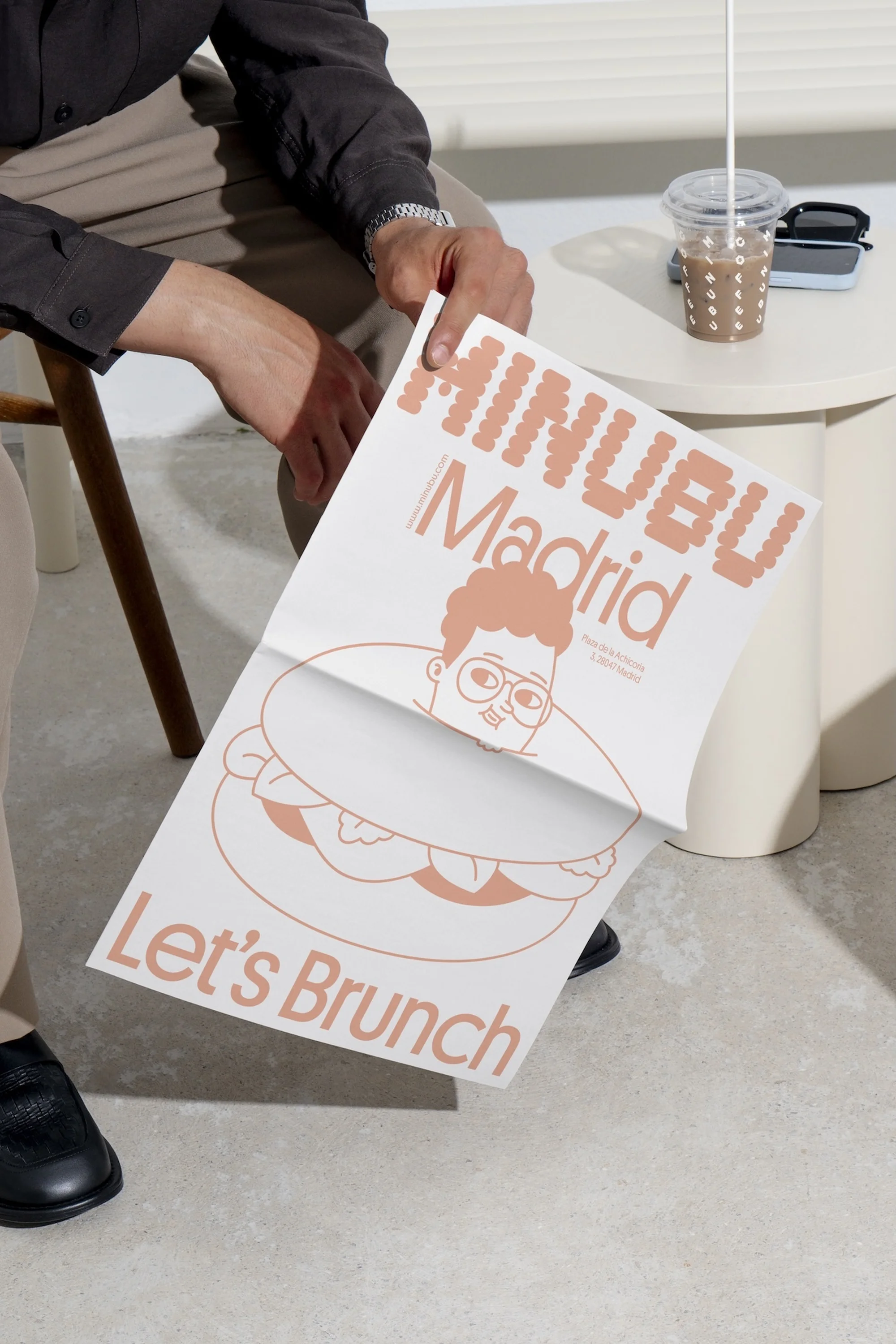 Person holds poster with cartoon graphic of a person inside a donut, next to table with drink and phone indoors, studio lighting.