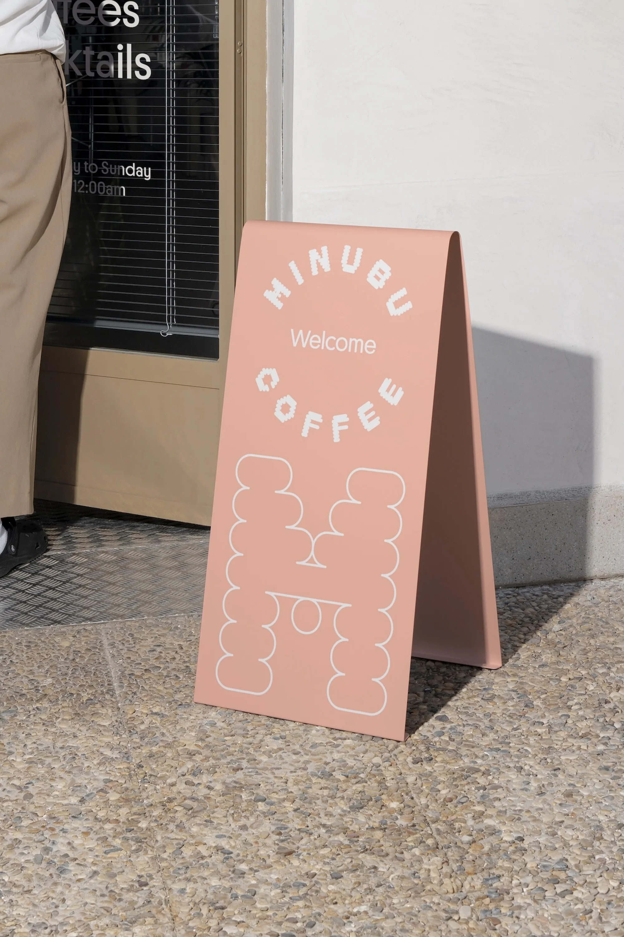 A-frame sign outside a coffee shop with 'Minubu Coffee Welcome' in stylized white text on a pink background and a person partially visible to the left.