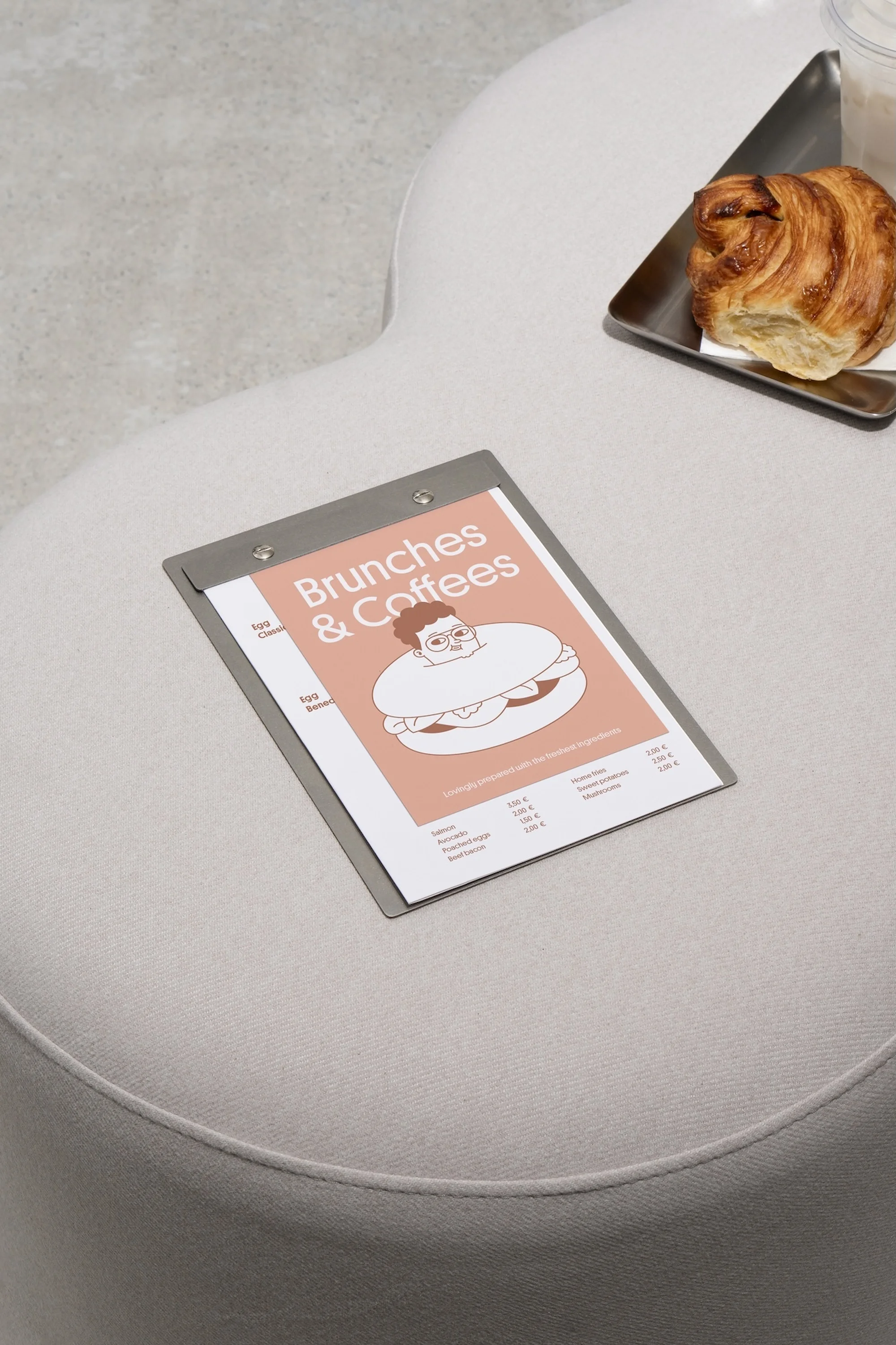 Overhead view of croissant and clipboard with a graphic of a person inside a sandwich sitting on an off-white padded surface.