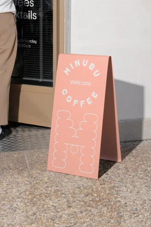 Pink sandwich board sign with Minubu Coffee branding sitting on pavement outside a storefront.