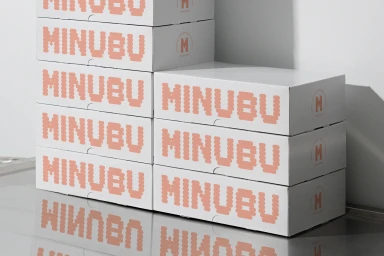 Stacked white Minubu Coffee boxes with pink pixelated typography on a reflective metal surface against a white wall.