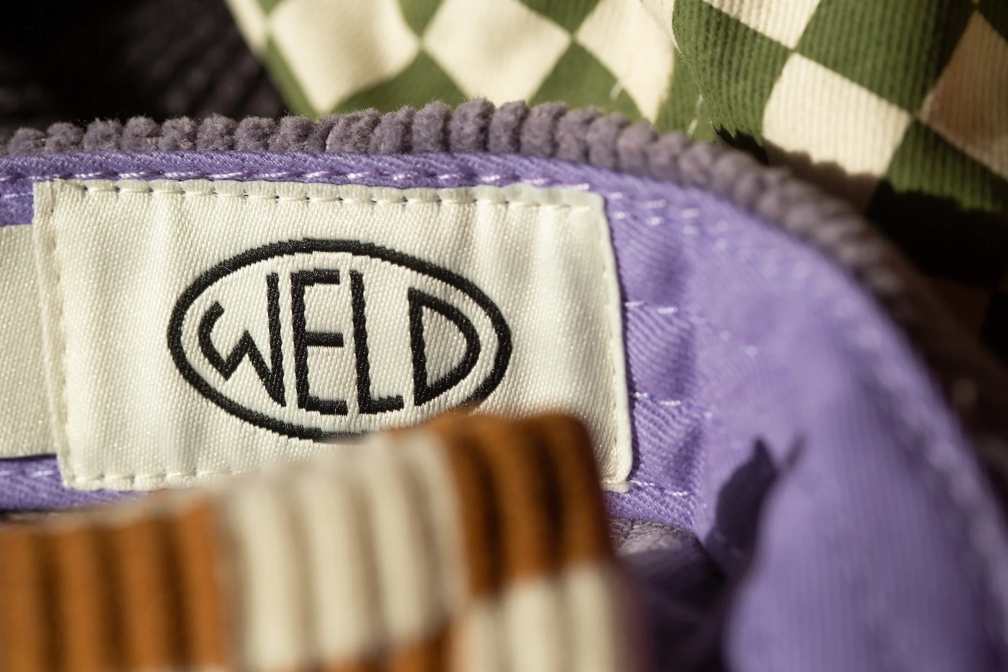 Close-up of a white clothing label with the word 'WELD' in black letters, attached to a light purple corduroy garment.
