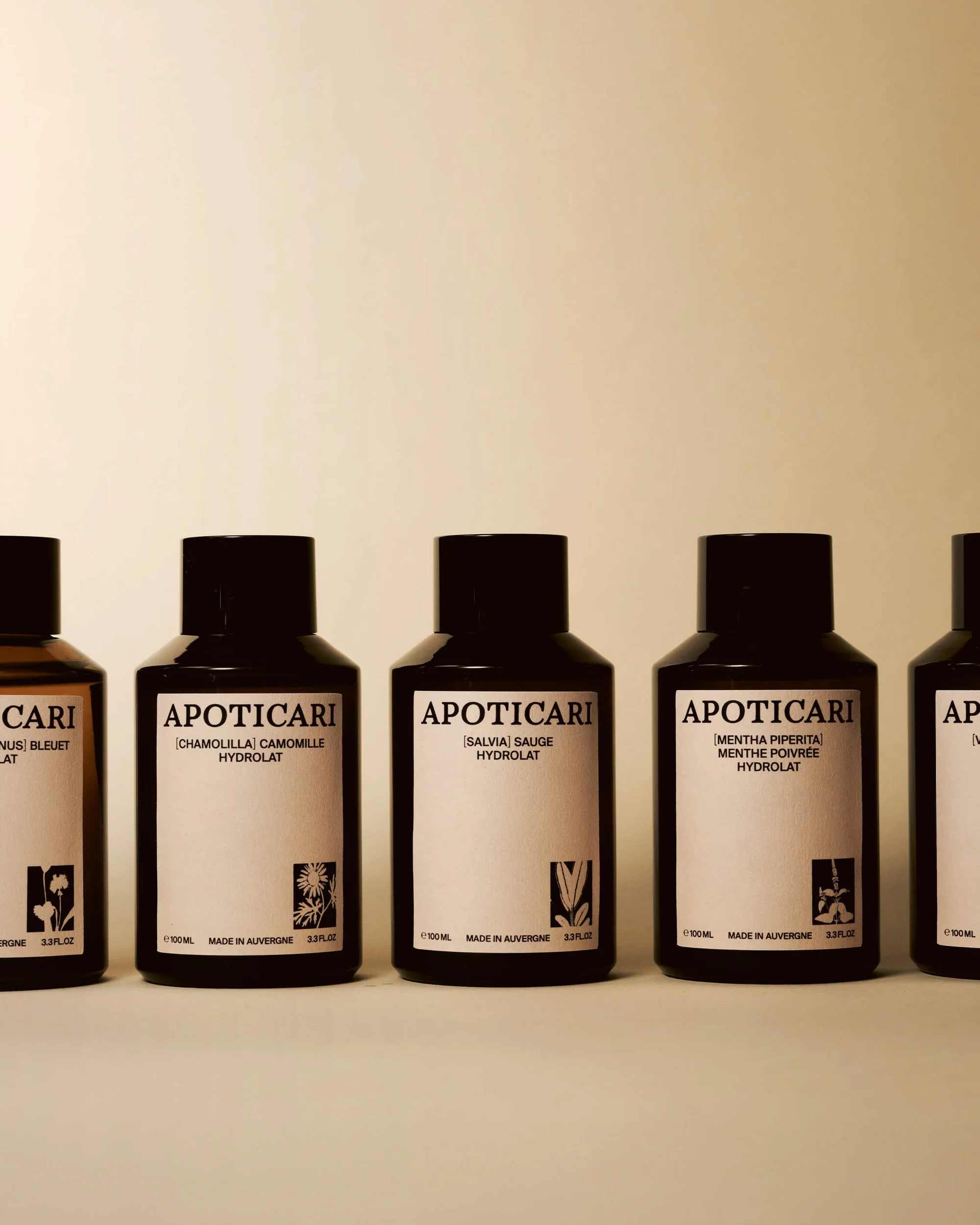 Five dark brown bottles of Apoticari Hydrolat skincare product are lined up on a white surface against a neutral background.