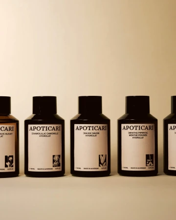 Five dark brown bottles of Apoticari Hydrolat skincare product are lined up on a white surface against a neutral background.