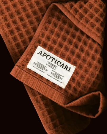 Close-up of a textured waffle weave towel in a warm brown tone, folded to reveal a white fabric label with black text, set against a dark background.