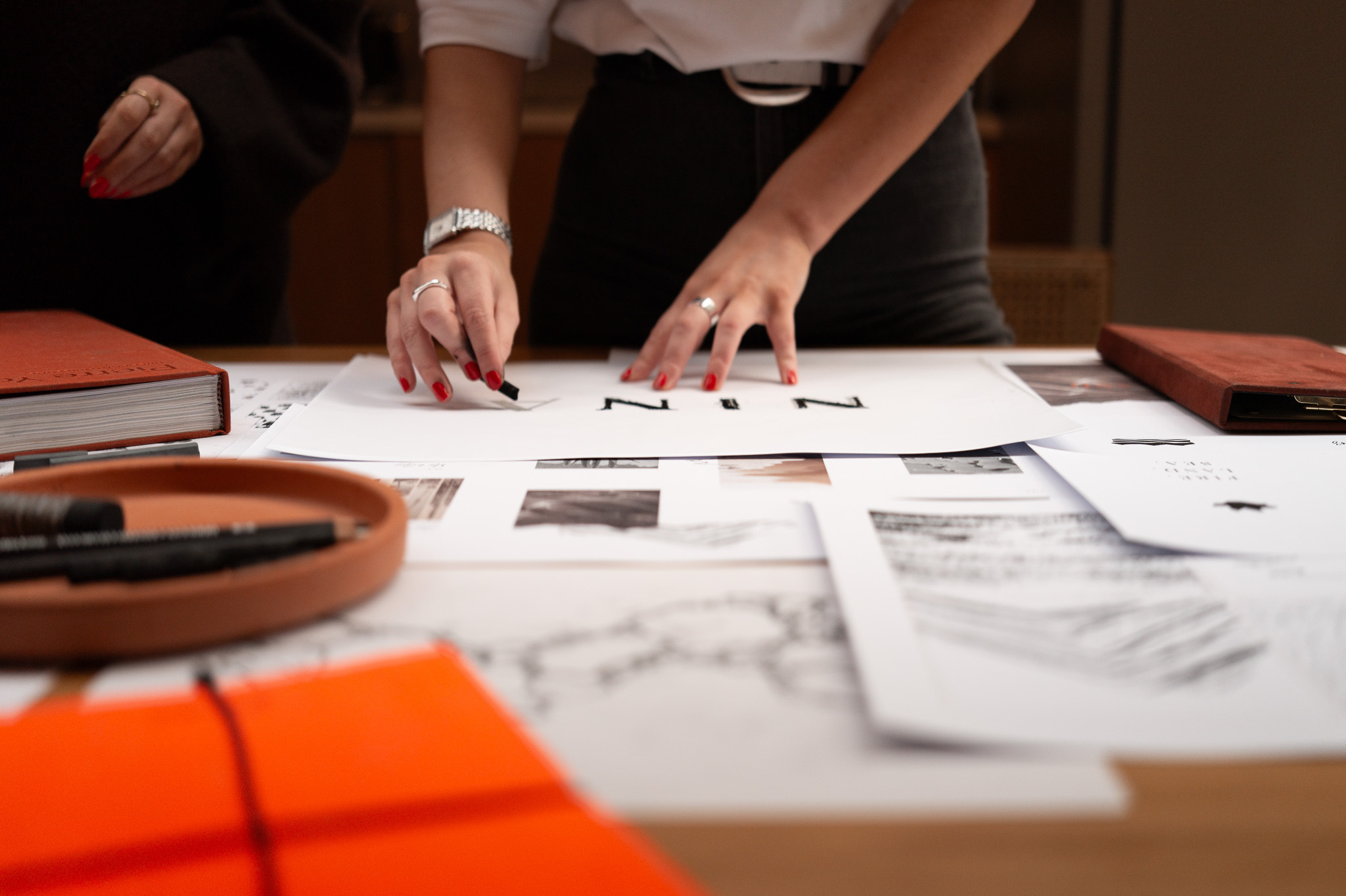 Two women at a table with art and design materials, working on a design or art project, drawing on paper with orange and brown accents.