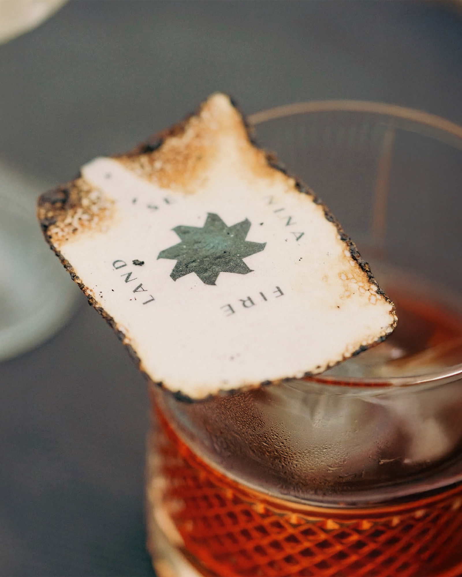 Close-up of a cocktail in a glass with an ornate label on top, displaying text and a star, with a textured surface visible.