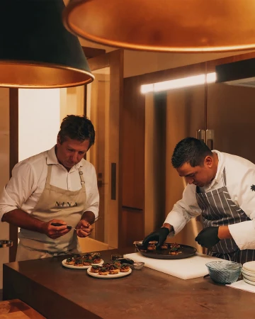 Two chefs in a restaurant kitchen plating appetizers, utilizing a wooden countertop and bathed in warm, inviting lighting from above.