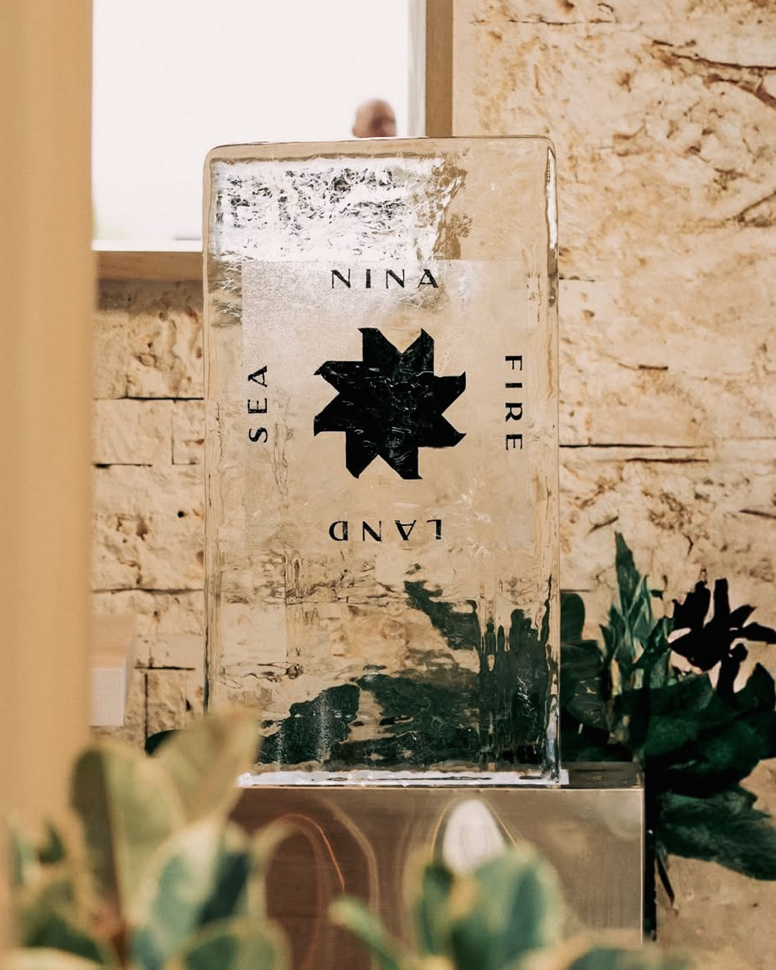Close-up of a clear ice sculpture with a black star design and the words NINA, SEA, LAND, and FIRE, with green plants nearby.