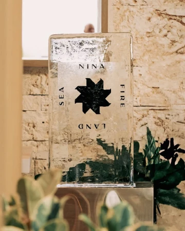 Close-up of a clear ice sculpture with a black star design and the words NINA, SEA, LAND, and FIRE, with green plants nearby.
