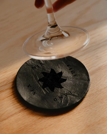 Close-up of a wine glass over a round black leather coaster with an eight-pointed star and inscriptions of 'Sea', 'Fire', 'Land', 'Wind'.