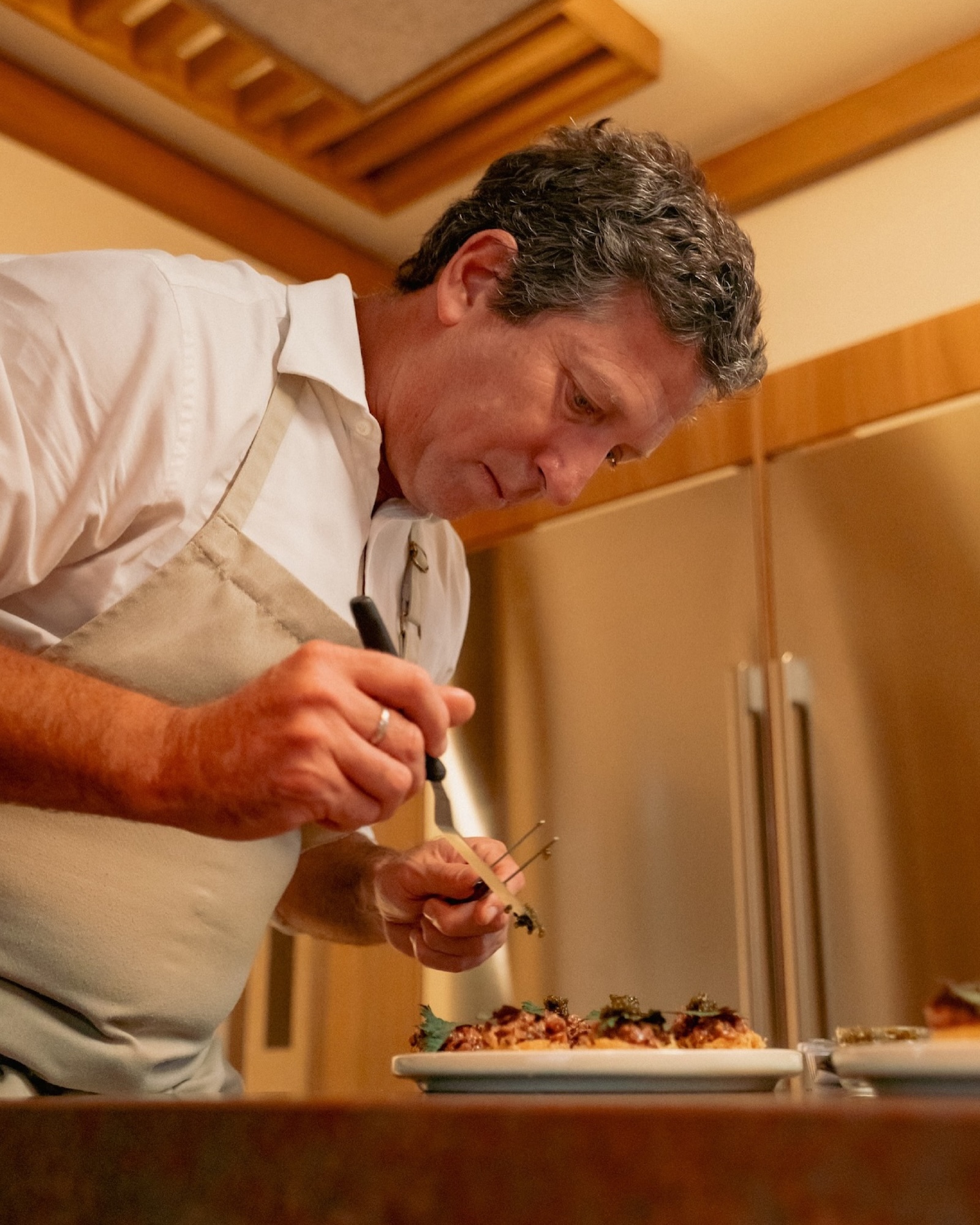 A chef in a white coat and beige apron is carefully plating food in a kitchen setting with stainless steel appliances and wooden elements.