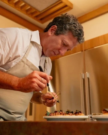 A chef in a white coat and beige apron is carefully plating food in a kitchen setting with stainless steel appliances and wooden elements.