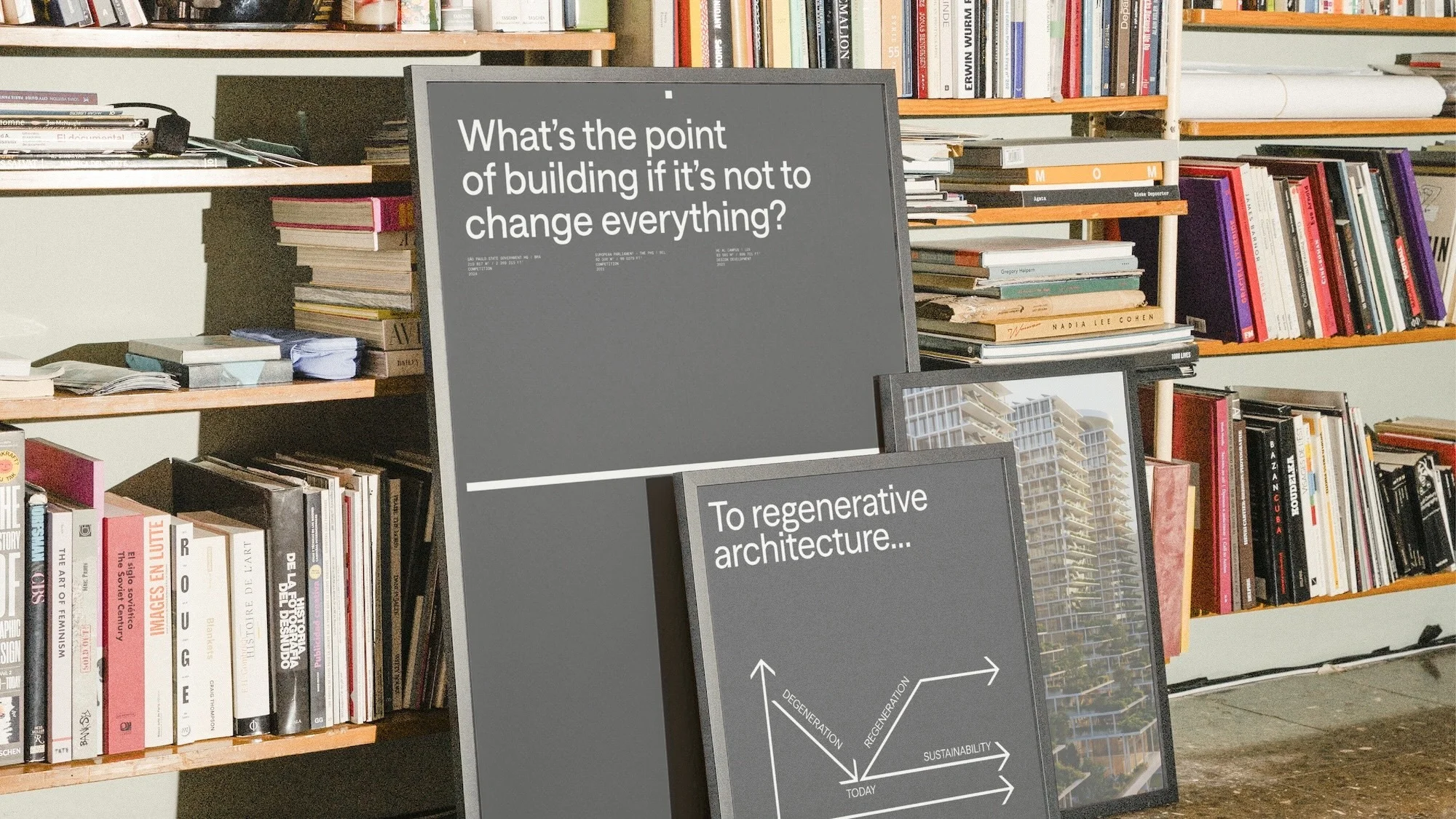 Two framed posters with architectural text and building imagery are leaning against a bookshelf filled with books in an indoor setting.
