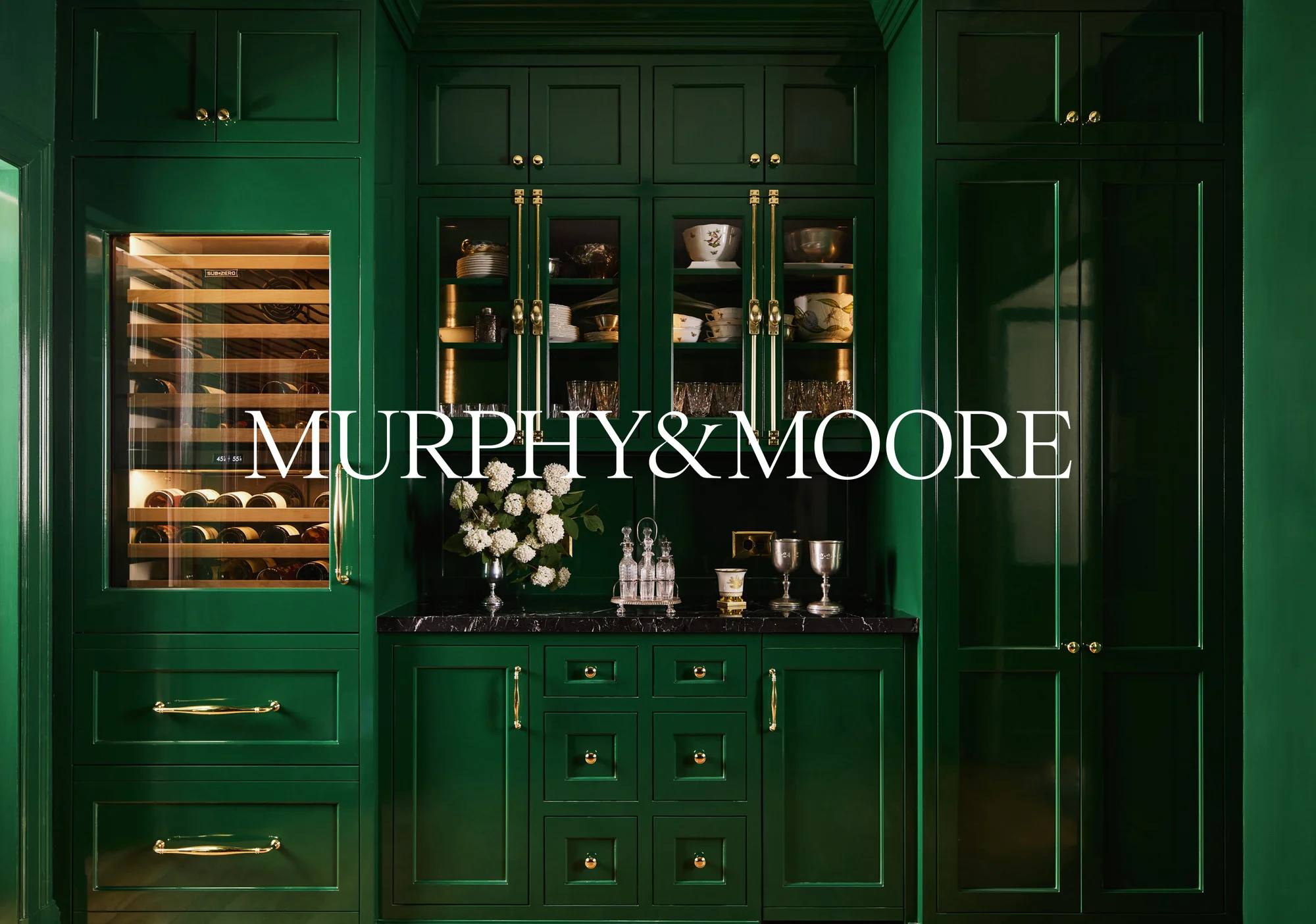 Elegant green kitchen with custom cabinetry, marble countertop, wine storage, and decorative dishware and barware.